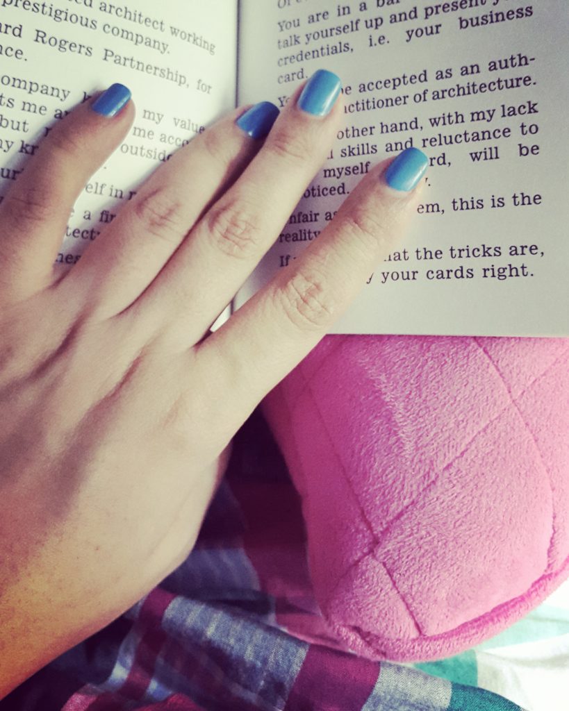 A picture of a girl's hand with blue nail color, holding a book on a pink pillow. 