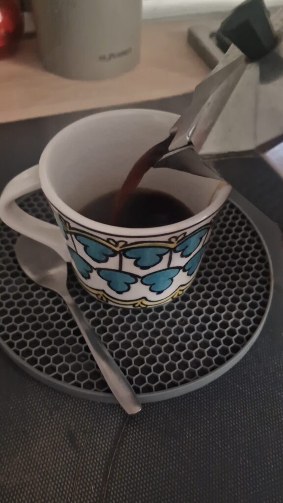 Picture of a moka pot pouring coffee into a white coffee cup. 